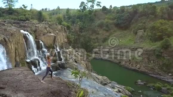 女孩在岩石上做瑜伽在瀑布从悬崖上流下来视频的预览图