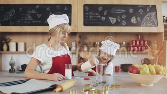 妈妈和女儿在一个舒适的厨房里一个小女儿把牛奶倒进玻璃杯里早餐烹饪概念视频的预览图
