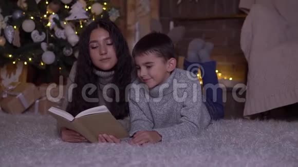 留着长发的妈妈给儿子读一本书并在圣诞节的时候亲吻他躺在圣诞树附近的地板上高清高清视频的预览图