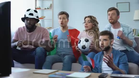 多种族的意大利球迷坐在沙发上观看比赛庆祝进球视频的预览图