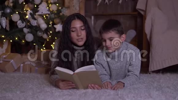 留着长发的妈妈给儿子读一本书并在圣诞节的时候亲吻他躺在圣诞树附近的地板上高清高清视频的预览图