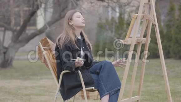 漂亮的年轻吸烟女孩坐在后院的户外画布上画画充满激情的成功艺术家视频的预览图