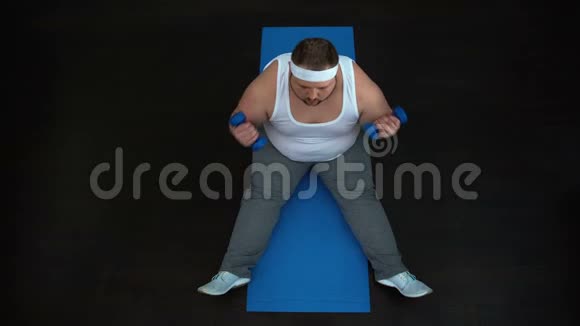 胖乎乎的男人坐在垫子上做二头肌卷发哑铃抽着手臂肌肉视频的预览图