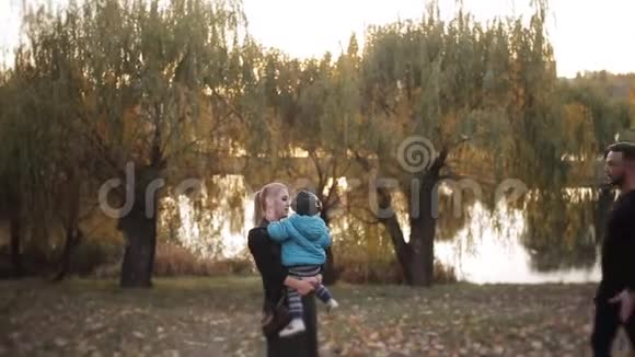 年轻的幸福家庭和小儿子在秋天公园散步日落时在公园附近的湖视频的预览图