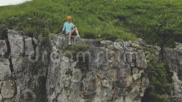 年轻的时髦女人坐在高高的岩石墙上的边缘空中观景无人机慢悠悠地飞行视频的预览图