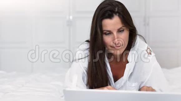喜欢在家玩得开心的女人躺在床上微笑着用笔记本电脑视频的预览图