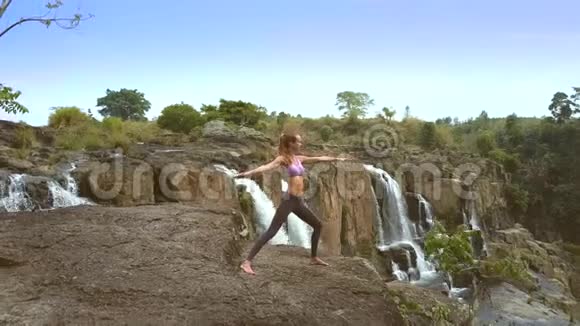 金发女郎站在瀑布旁平坦的岩石上做瑜伽姿势视频的预览图