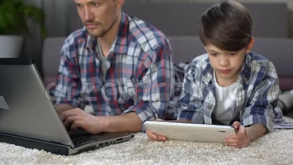 小儿子在家里的地板上玩平板电脑而爸爸在用笔记本电脑视频的预览图