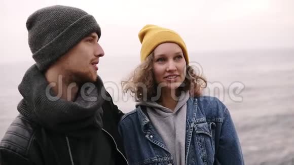 一对年轻的幸福夫妇站在一起在海边拥抱穿暖和的衣服戴冷帽的年轻人视频的预览图