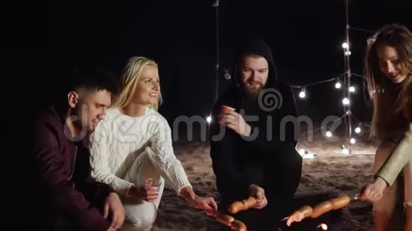 傍晚海滩野餐时四个年轻朋友在篝火旁串烧香肠视频的预览图