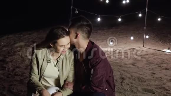 在海滩上浪漫的夜晚快乐的女孩和男人坐在沙滩上背景是装饰的灯光视频的预览图