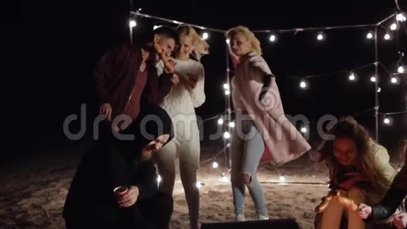 篝火晚会朋友们在沙滩上的灯光下在串串上享用饮料和香肠视频的预览图