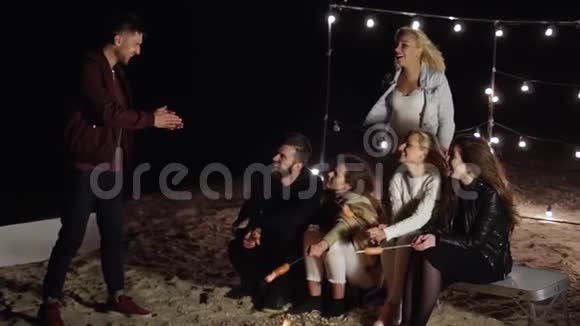快乐的朋友在夜晚的海滩上玩游戏野餐时在露天的烤肉串上放香肠视频的预览图