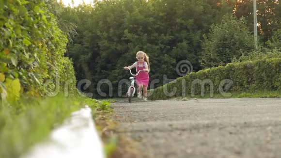 一个快乐美丽金发碧眼的小女孩穿着粉红色的裙子跳投骑着一辆儿童自行车上路视频的预览图