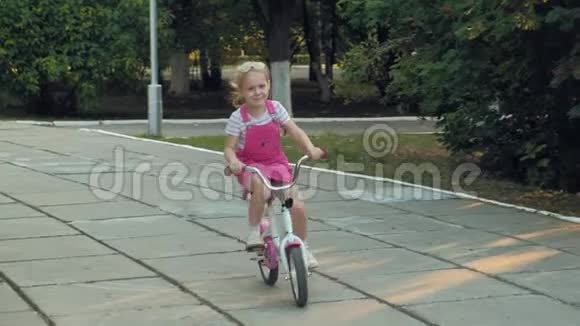 一个快乐美丽金发碧眼的小女孩穿着粉红色的裙子跳投骑着一辆儿童自行车上路视频的预览图