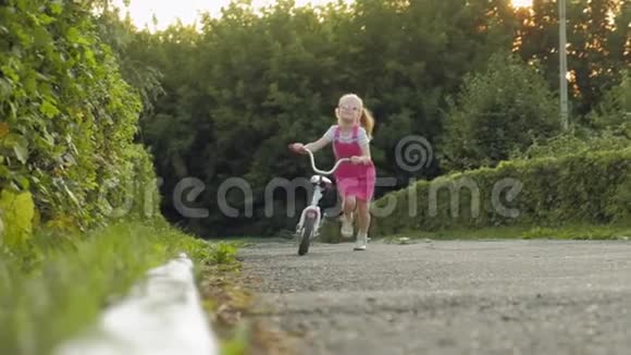 一个快乐美丽金发碧眼的小女孩穿着粉红色的裙子跳投骑着一辆儿童自行车上路视频的预览图