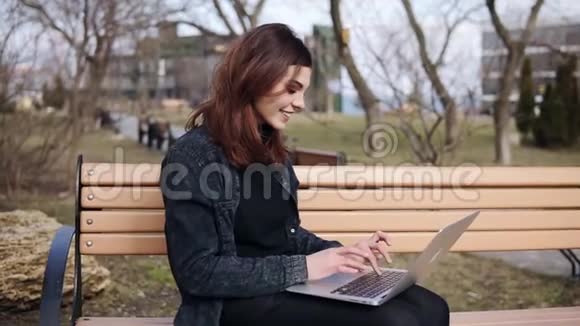 20岁姐姐坐在公园的长凳上在她的笔记本电脑键盘上打字很有魅力享受免费时间视频的预览图