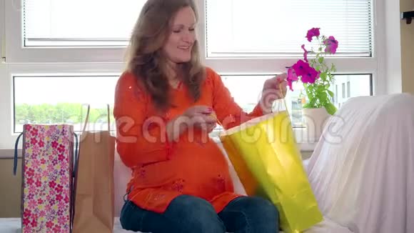 快乐的孕妇在买完东西后坐在沙发上看婴儿衣服视频的预览图