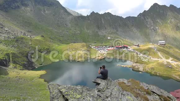 坐在岩石上的年轻人拍摄被群山环绕的湖的照片视频的预览图