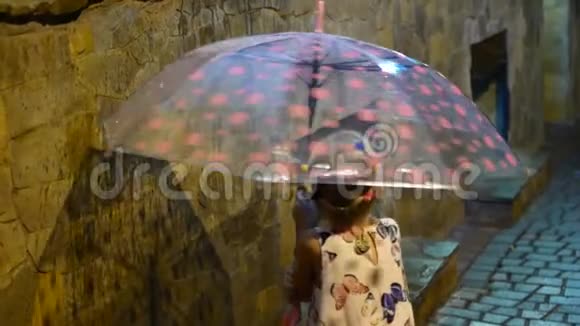 一个可爱的小女孩坐在水坑旁边雨滴顺着一把多色的伞流下来孩子微笑着视频的预览图