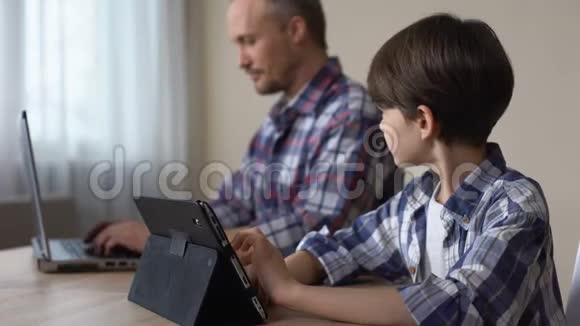 男孩子在平板电脑上玩游戏而父亲则在家里做笔记本电脑视频的预览图