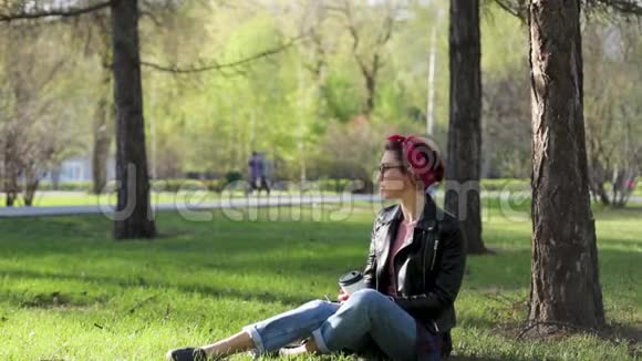 美丽年轻时尚的女人坐在公园的一棵树下喝着一杯咖啡大自然中的外卖咖啡时尚时尚时尚视频的预览图