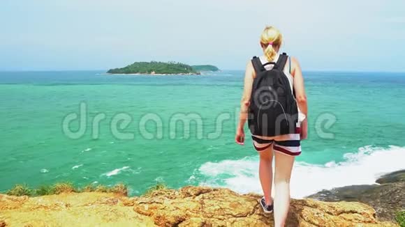 一个背着背包的旅行者女孩站在沙滩上望着远方有岩石和大海的美丽海景视频的预览图