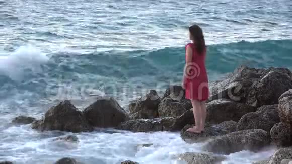 美丽的年轻女子在岩石海岸张开双臂在风中浪花飞溅视频的预览图
