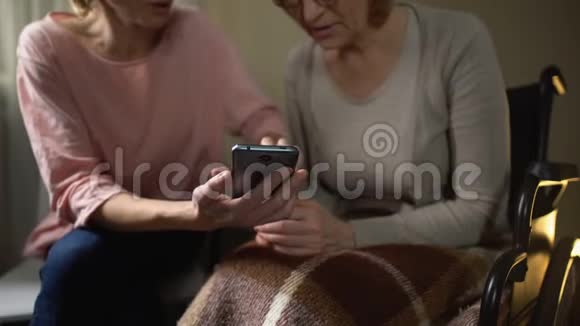 善良的女性志愿者向轮椅上的老妇人展示如何使用智能手机视频的预览图