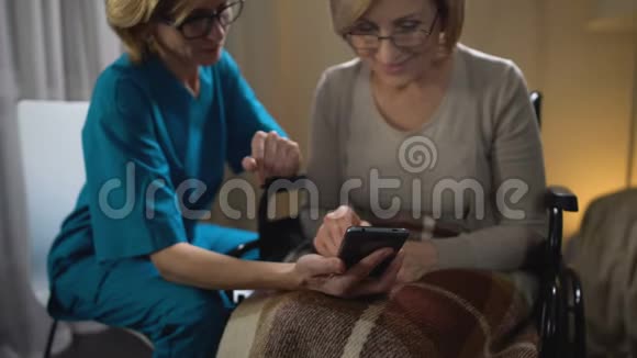友好的志愿者教坐轮椅的妇女使用智能手机养老院视频的预览图