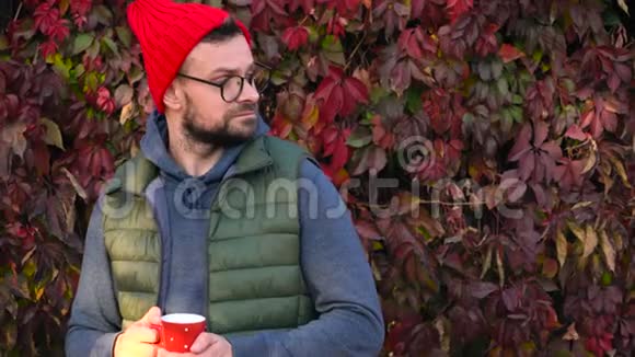 戴着红帽子手里拿着杯子的人正在户外喝茶或咖啡在温暖的秋日里用智能手机视频的预览图