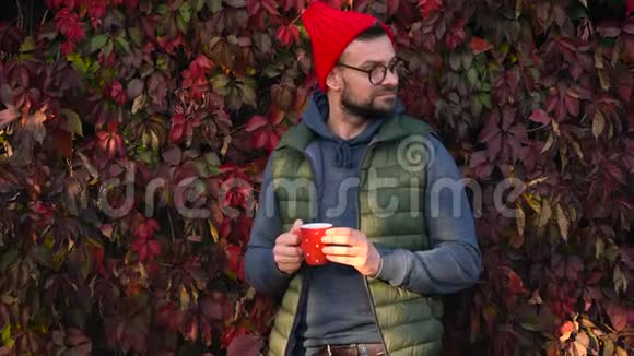 戴着红帽子手里拿着杯子的人正在户外喝茶或咖啡在温暖的秋日里用智能手机视频的预览图