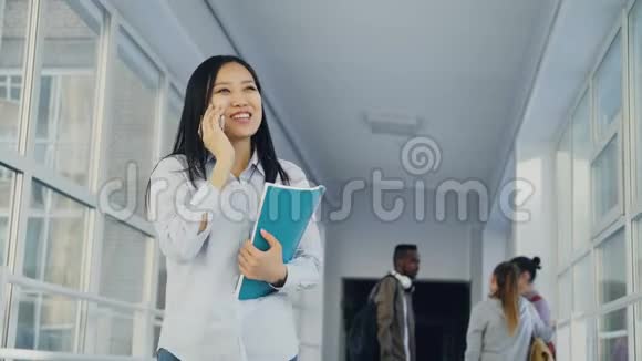 年轻美丽的亚裔女学生在电话中微笑着站在大学里的画像视频的预览图