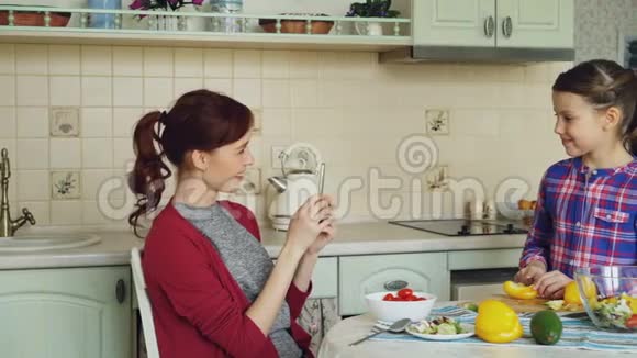 年轻的母亲在智能手机摄像头上谈论她可爱的微笑女儿在厨房切菜的照片视频的预览图
