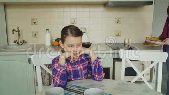 年轻的母亲带着美味的松饼和羊角面包给她可爱的女儿她坐在厨房的桌子旁等着视频的预览图