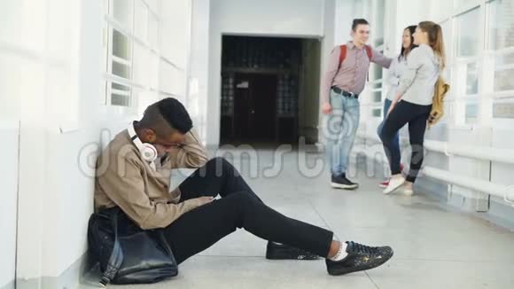 在大学走廊里坐在地板上看着不同的侧面时令严肃的非裔美国男学生心烦意乱视频的预览图