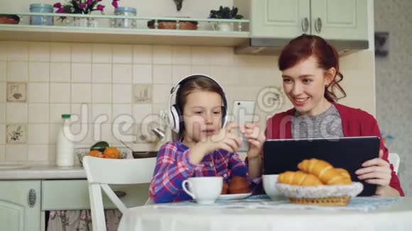 快乐的母亲和可爱的女儿早上在家厨房吃早餐小女孩在玩手机视频的预览图