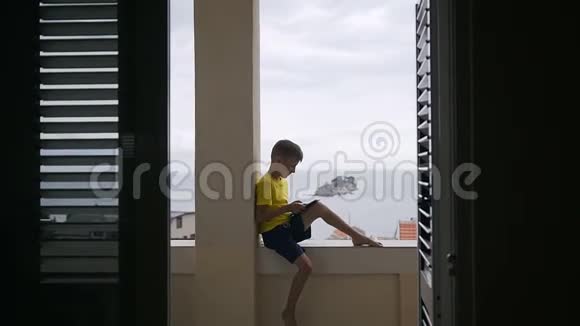 一个小男孩用他的平板电脑玩游戏而坐在阳台在家里的海洋背景青少年玩视频的预览图