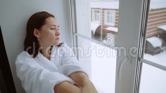 一个穿着白色外套的沮丧的女人坐在窗台上向外看着街道外面正在下雪一片悲伤视频的预览图