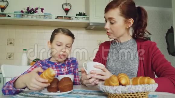 快乐的母亲和可爱的女儿吃早餐吃松饼和羊角面包在家里在现代厨房里聊天视频的预览图