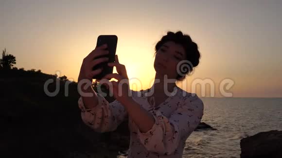 可爱的白种人布鲁内特女孩穿着轻装在日出的时候在海上方的悬崖上做自拍视频的预览图