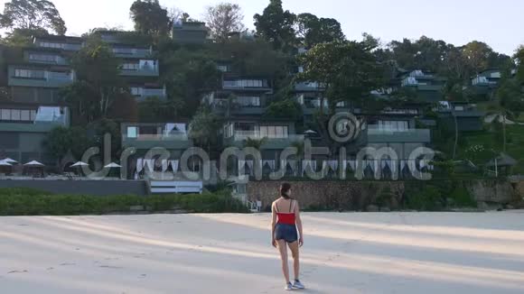 日出时美丽的红衣女郎在海滩边散步到豪华的温泉度假酒店视频的预览图