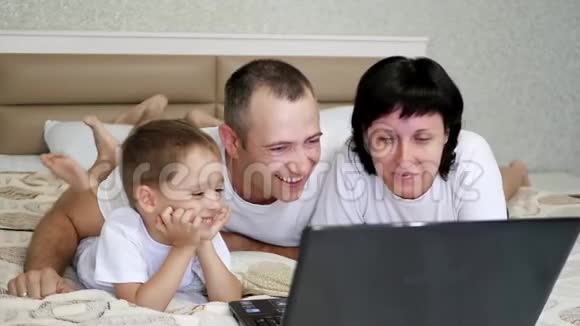 幸福的家庭爸爸妈妈和宝宝躺在床上看着笔记本电脑慢悠悠地笑着视频的预览图