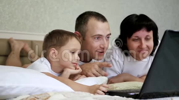 幸福家庭爸爸妈妈和孩子躺在床上看着笔记本电脑屏幕慢悠悠地笑着视频的预览图