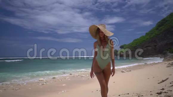 穿着绿松石比基尼和戴着一顶大草帽的漂亮成年女孩在热带海滩的沙滩上散步背景是视频的预览图