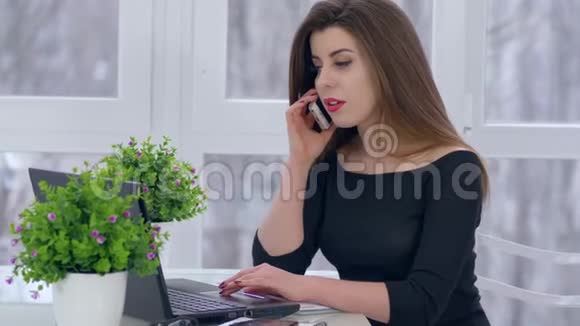 打电话年轻女人在智能手机上聊天在电脑上工作坐在房间的桌子上视频的预览图