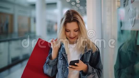 微笑的金发女孩在手机里输入一些东西带着购物袋在购物中心周围走动视频的预览图