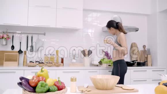 年轻的亚洲女人感到快乐的舞蹈听音乐喝橙汁在厨房做运动服装沙拉视频的预览图