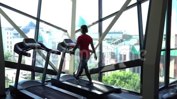 漂亮的白种人女孩在运动健身房的跑步机上跑步带电话和耳机相机在4K视频的预览图
