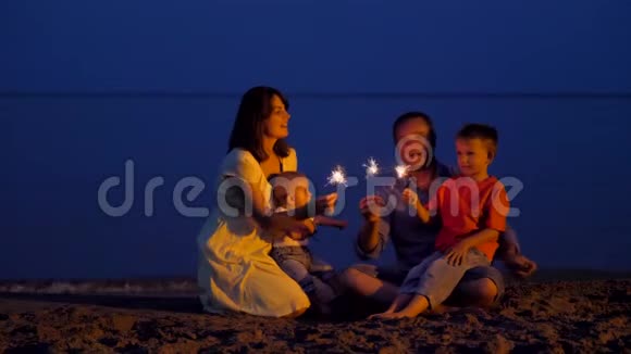 快乐的年轻家庭在黄昏的海滩上庆祝孩子们和父母在唱歌手捧烟花视频的预览图